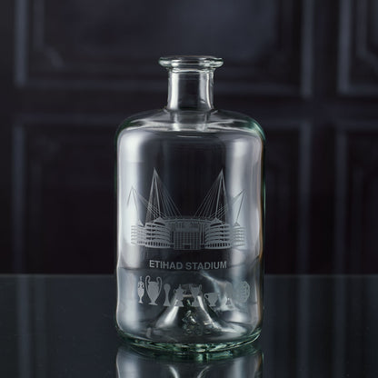 Apothecary-style glass decanter engraved with a line-art interpretation of the Etihad Stadium and trophy silhouettes, photographed on a dark background.