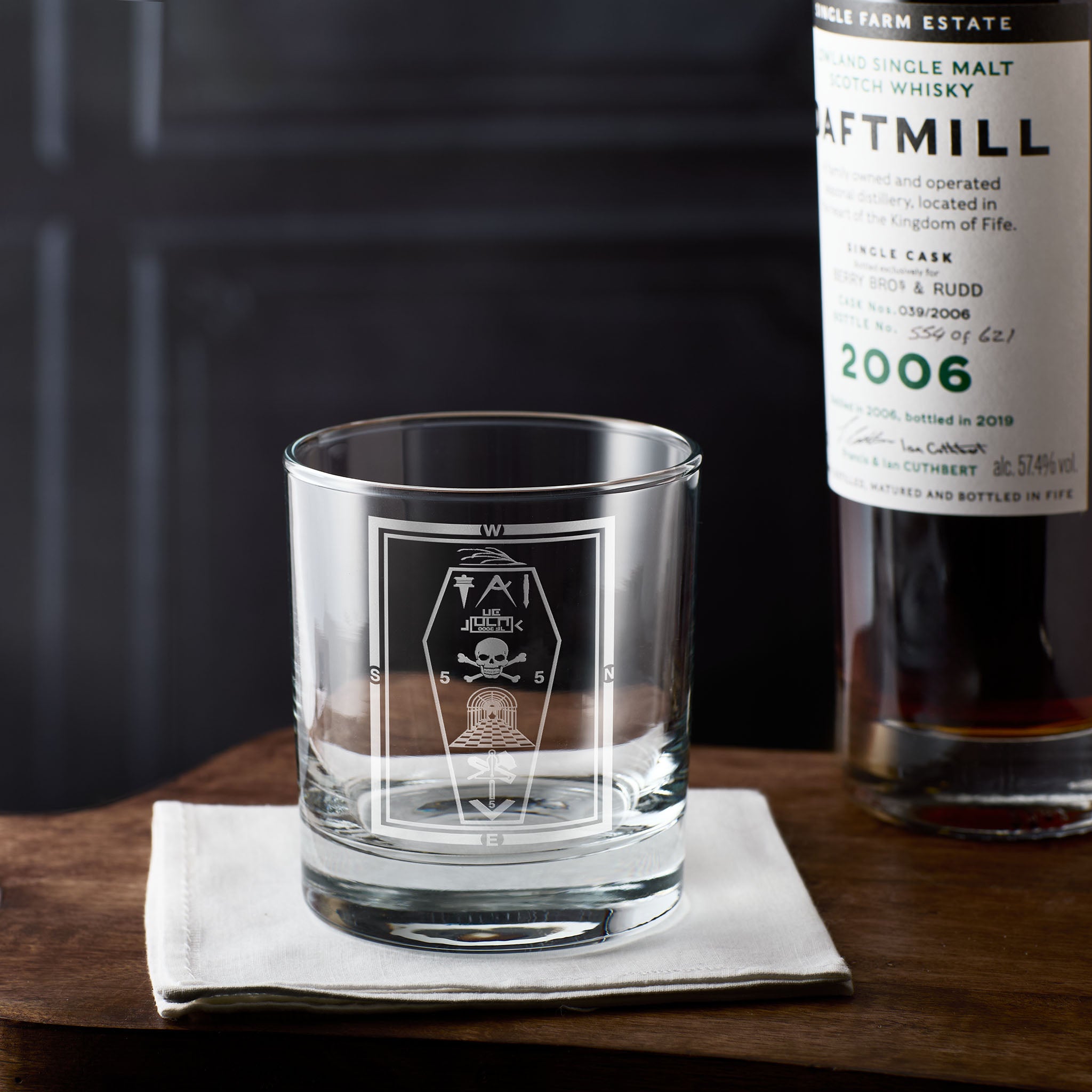 Whiskey glass with masonic 3rd degree tracing board and skull design on a white napkin next to a bottle of Daftmill whisky.