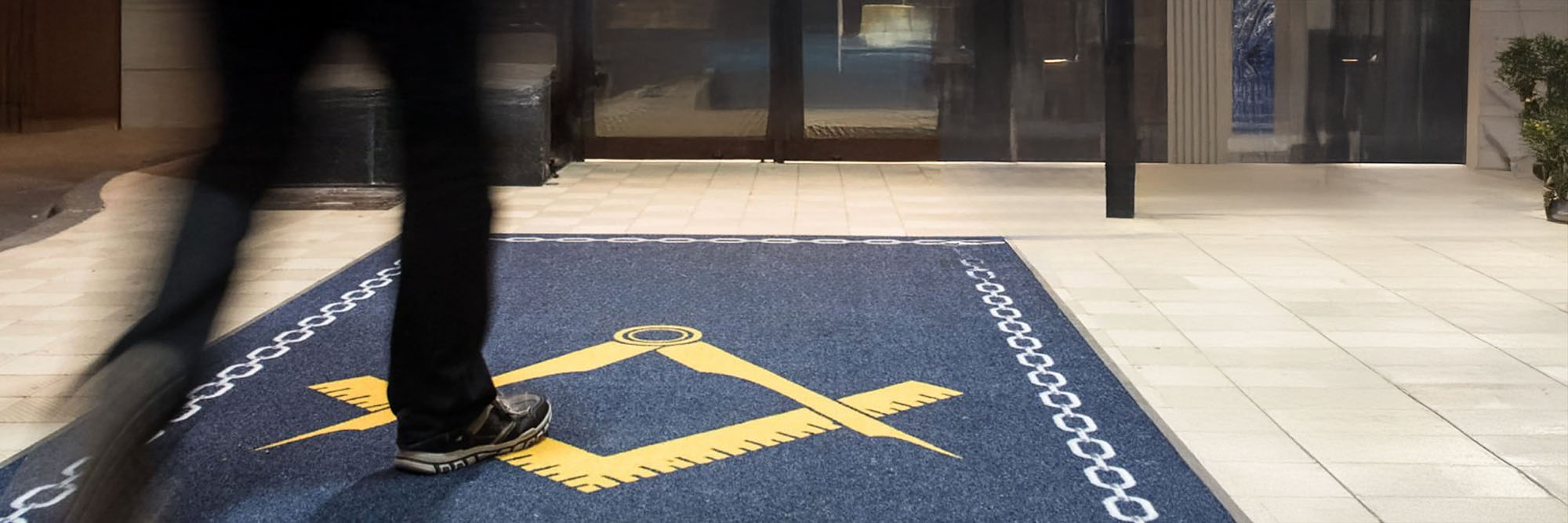 Person walking on a blue doormat with Masonic symbols at the entrance of a building.