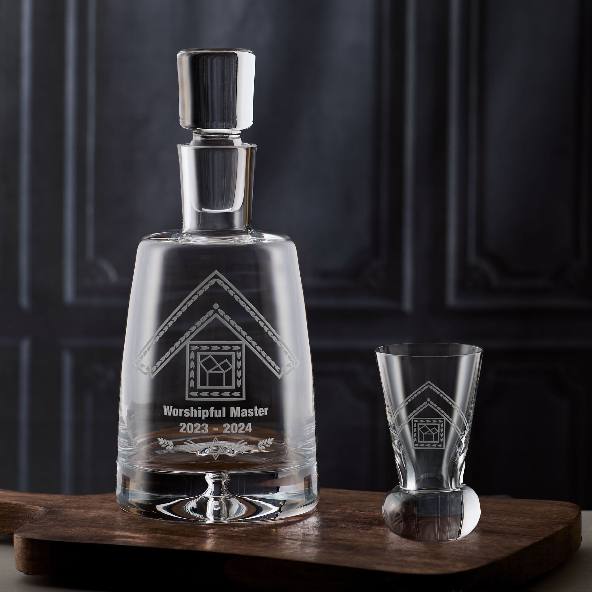 Masonic glass decanter and firing glass with engraved past master design on a wooden surface against a dark background