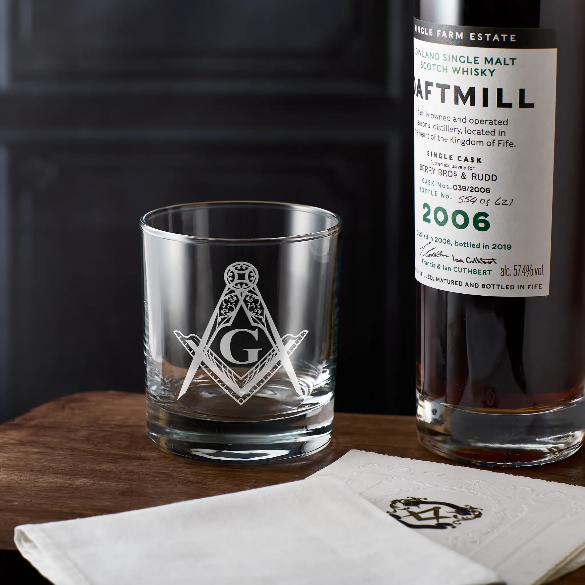 Whiskey glass with Masonic square and compass emblem next to a bottle of dafftamill whisky on a wooden surface.