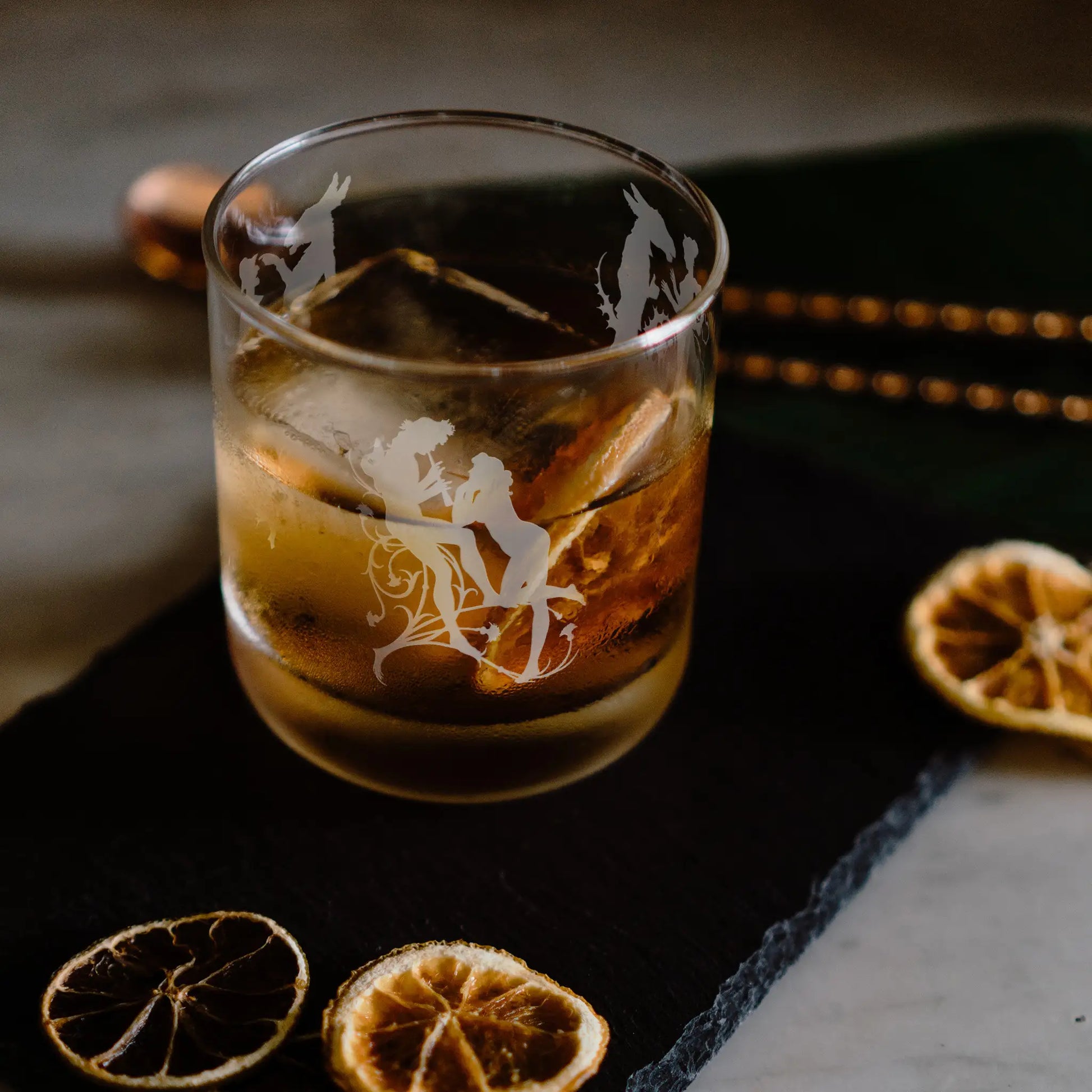 Engraved whiskey glass featuring Shakespear's Midsummers Nights Dream characters with ice and orange slices on a dark surface