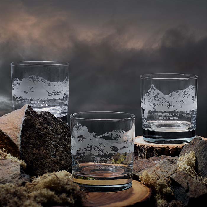 Three etched glass tumblers on a natural wooden surface with mountain landscapes.