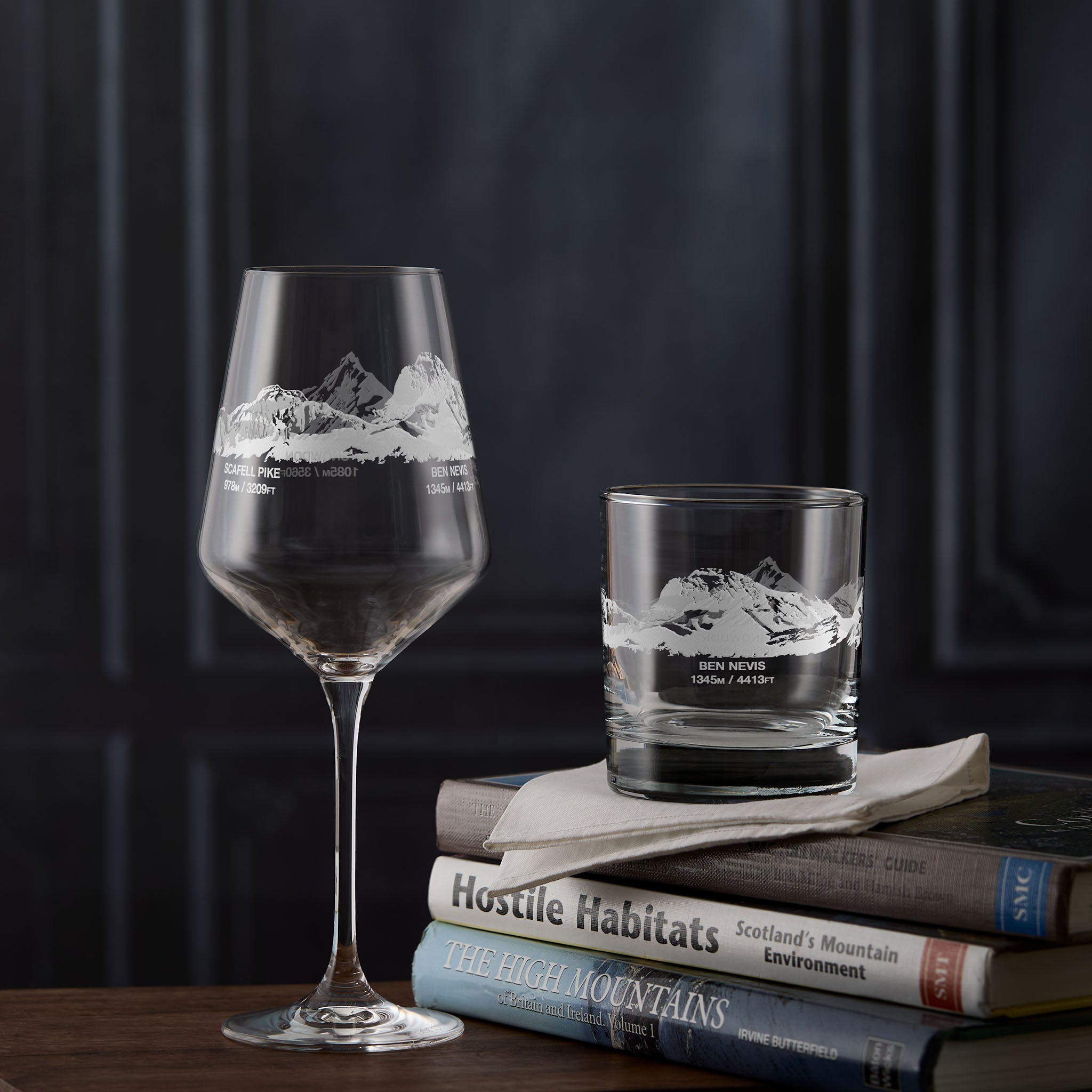 Two engraved glasses with UK mountain etching on a stack of books against a dark background