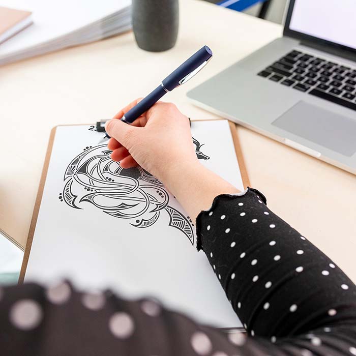 Person drawing on a notepad with a pen, sitting at a desk with a laptop.
