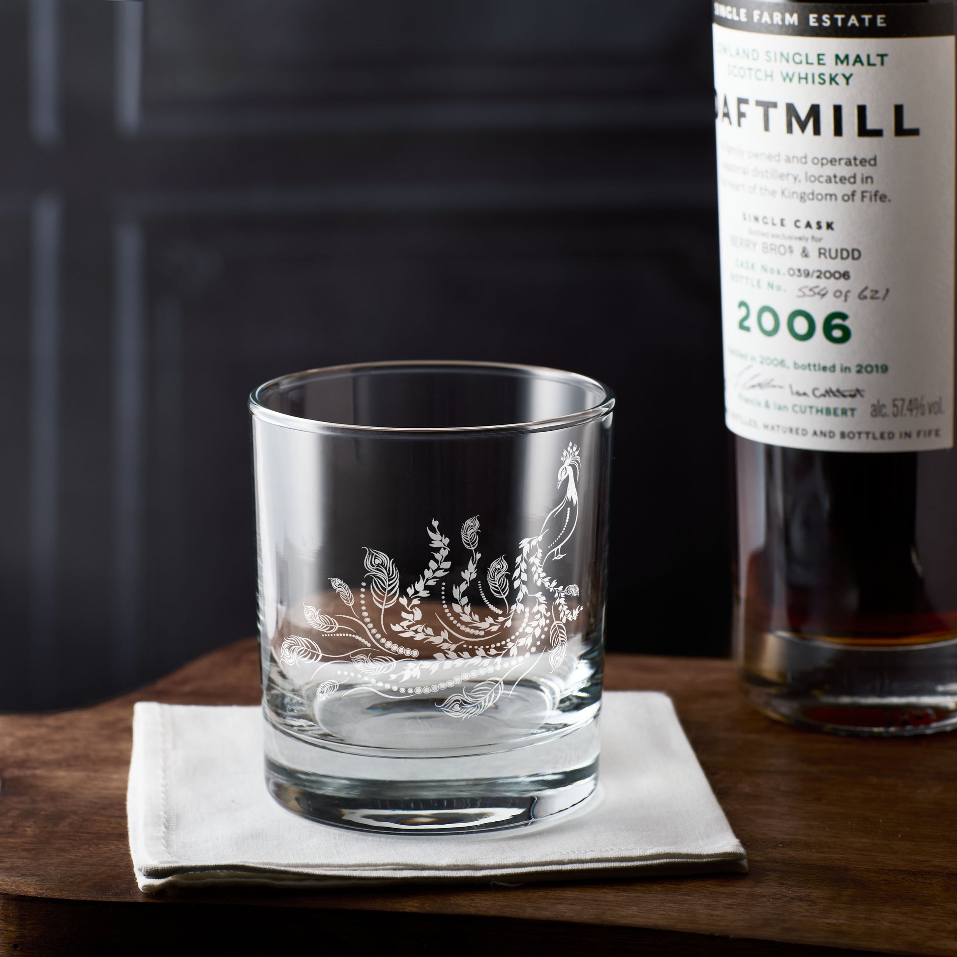 Engraved peacock on a whisky glass placed on a wooden surface with a bottle of Daftmill whisky in the background.