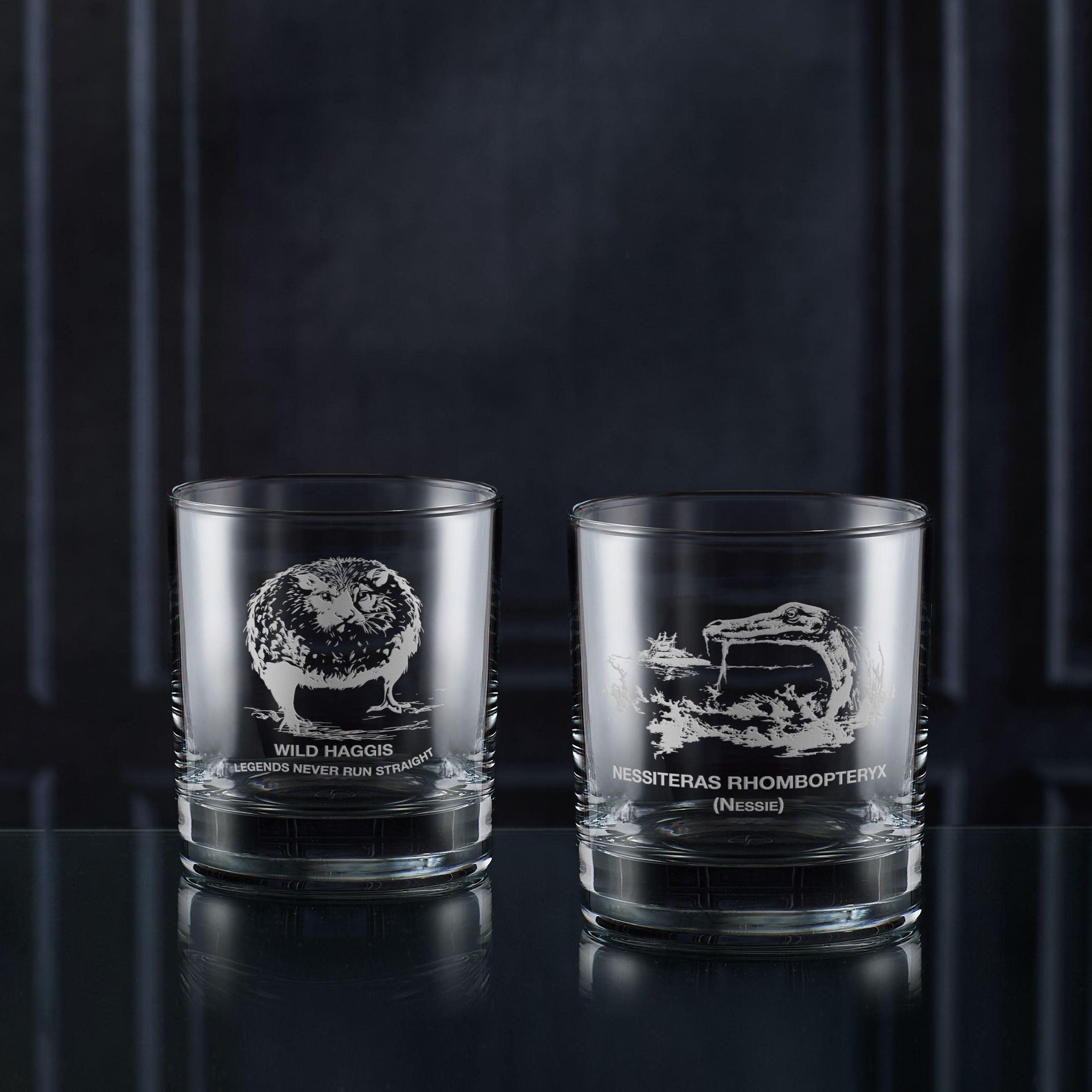Two glass whisky tumblers with engraved scottish myth designs and text on a dark background