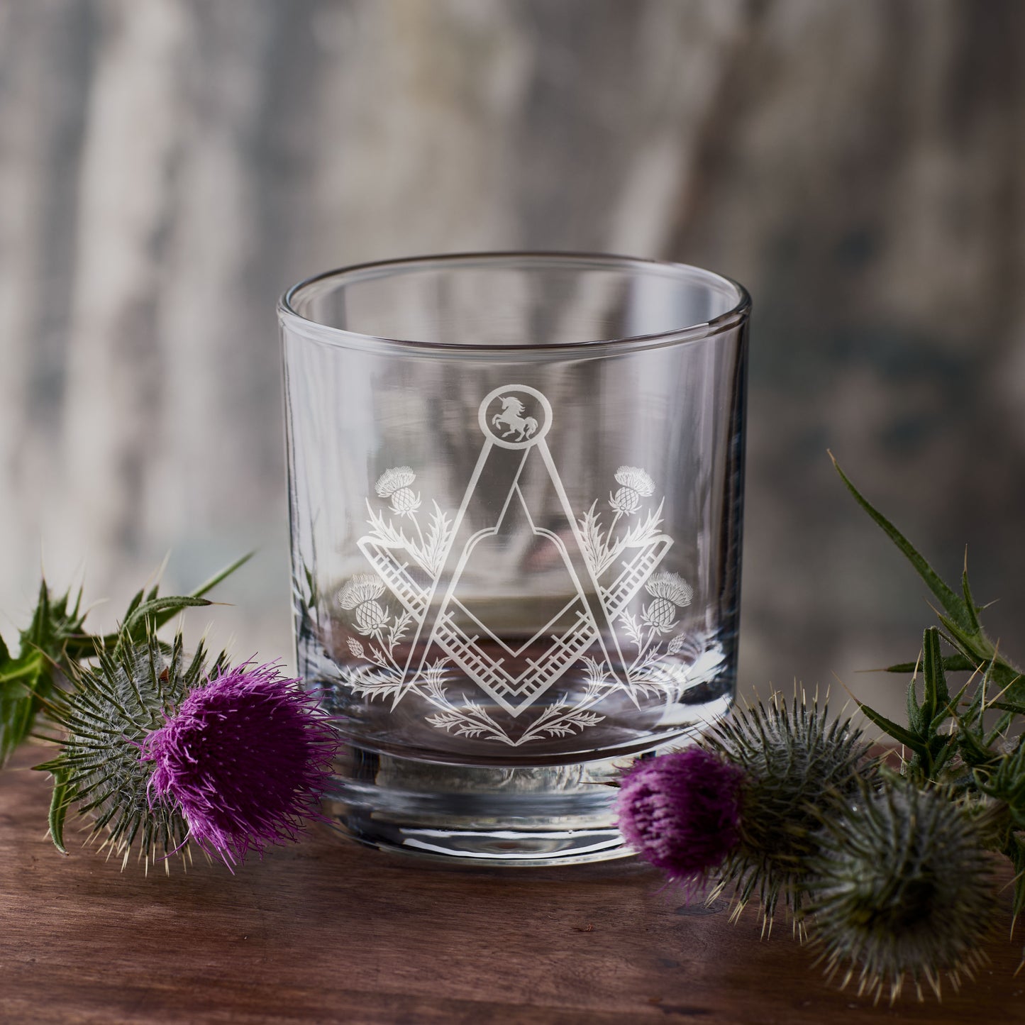 Clear glass with Masonic square and compass design surrounded by purple thistles on a wooden surface