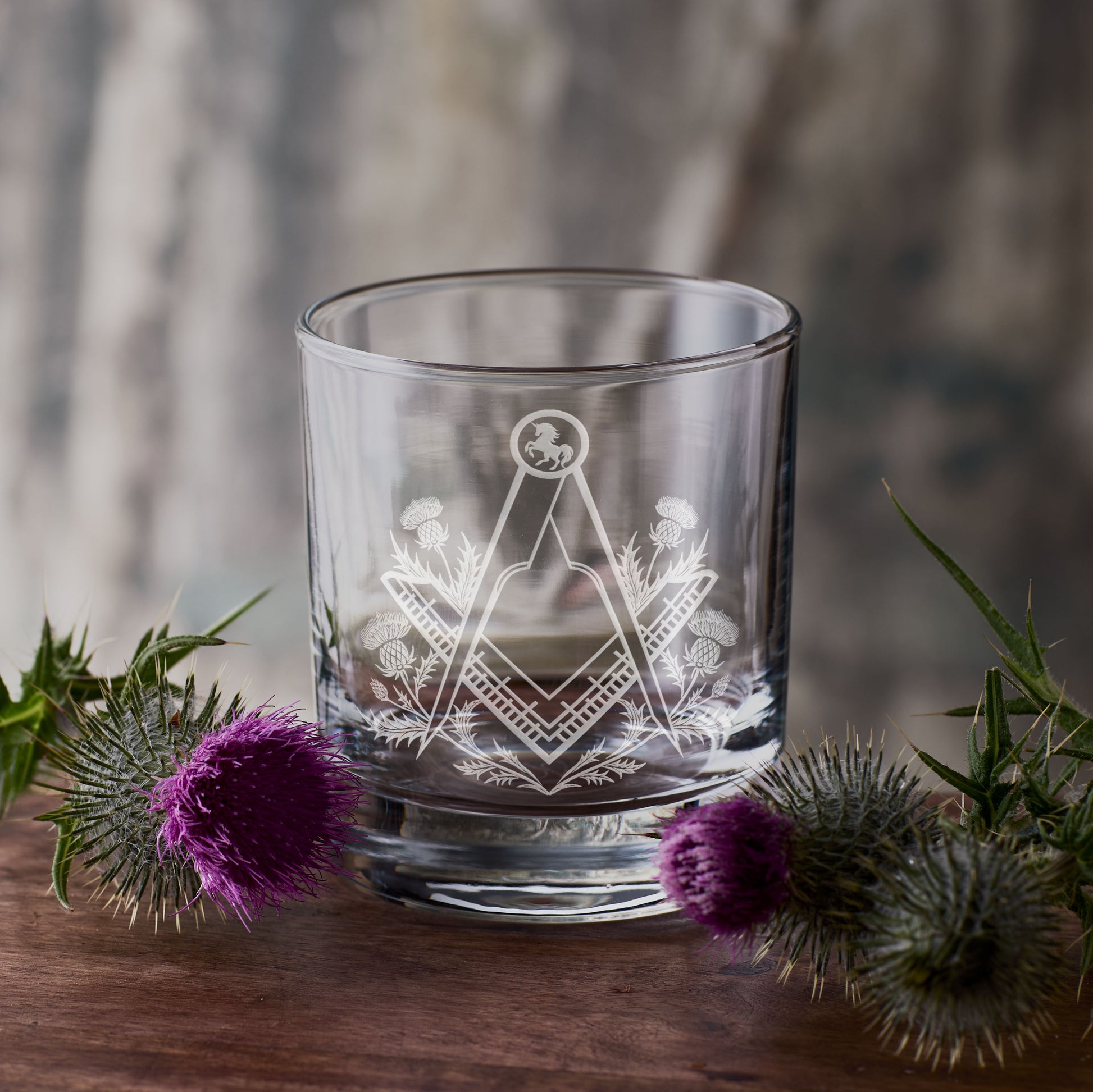 Clear glass with Masonic square and compass design surrounded by purple thistles on a wooden surface