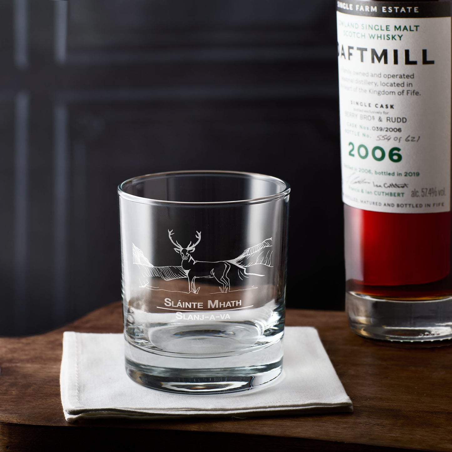 Whisky glass with engraved scottish stag design on a wooden surface next to a bottle of whisky.