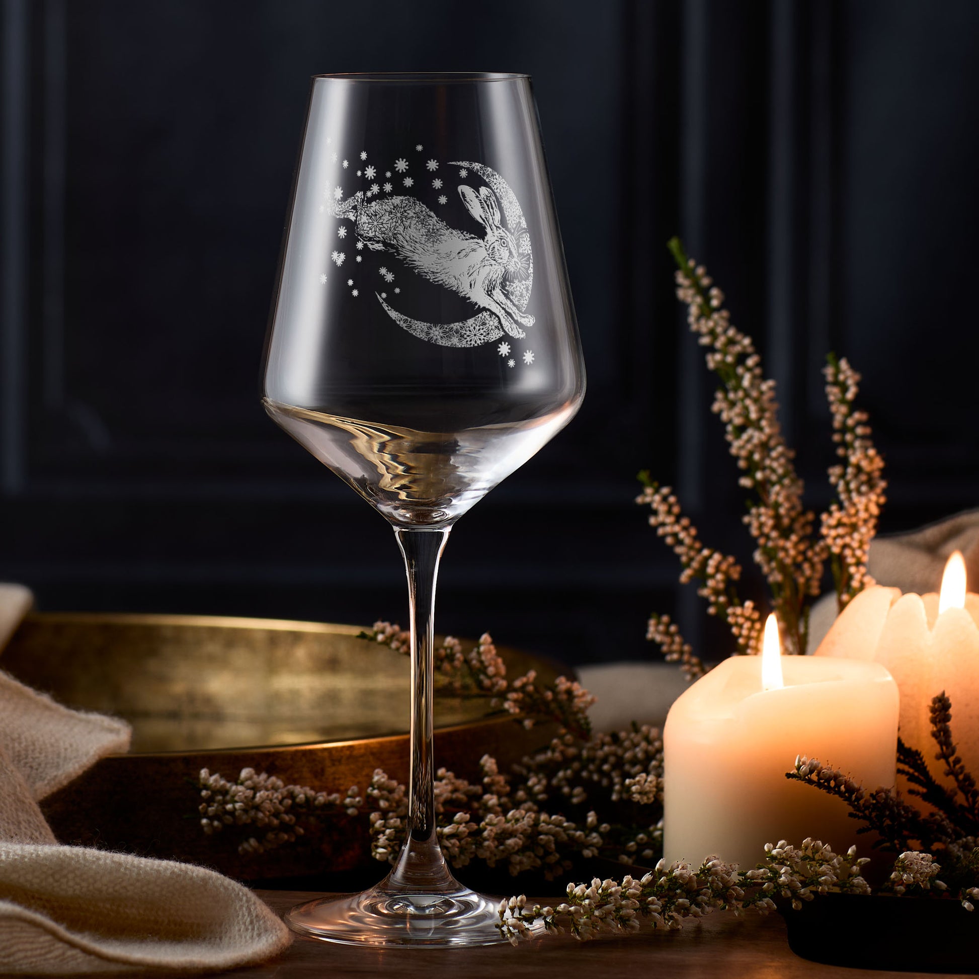 Wine glass with engraved design of a hare and moon, surrounded by candles and dried flowers on a dark background.