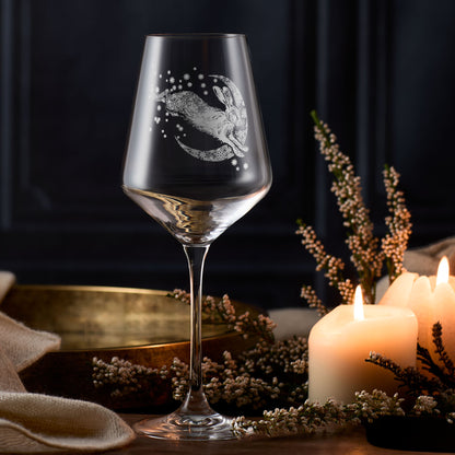 Wine glass with engraved design of a hare and moon, surrounded by candles and dried flowers on a dark background.