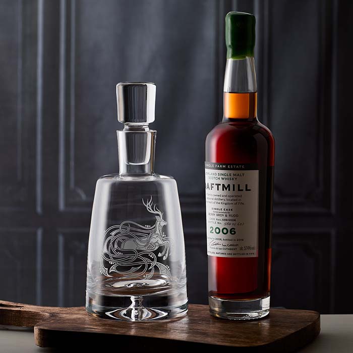 Clear glass decanter with engraved stag design next to a bottle of whisky on a wooden surface.