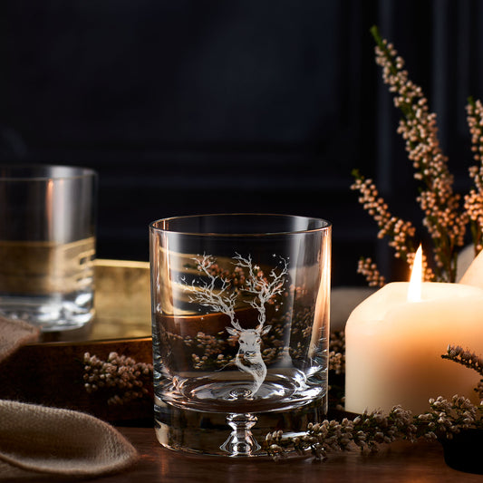 Whisky glass with an engraving of a deer, stars and winter branches placed on a table with a candle and decorative elements