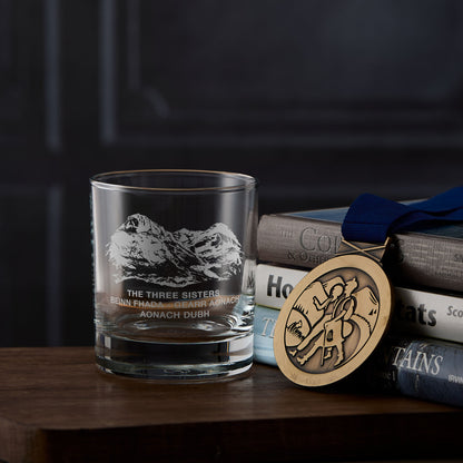 Glass with Glencoe mountain engraving, books, and a medal on a wooden surface
