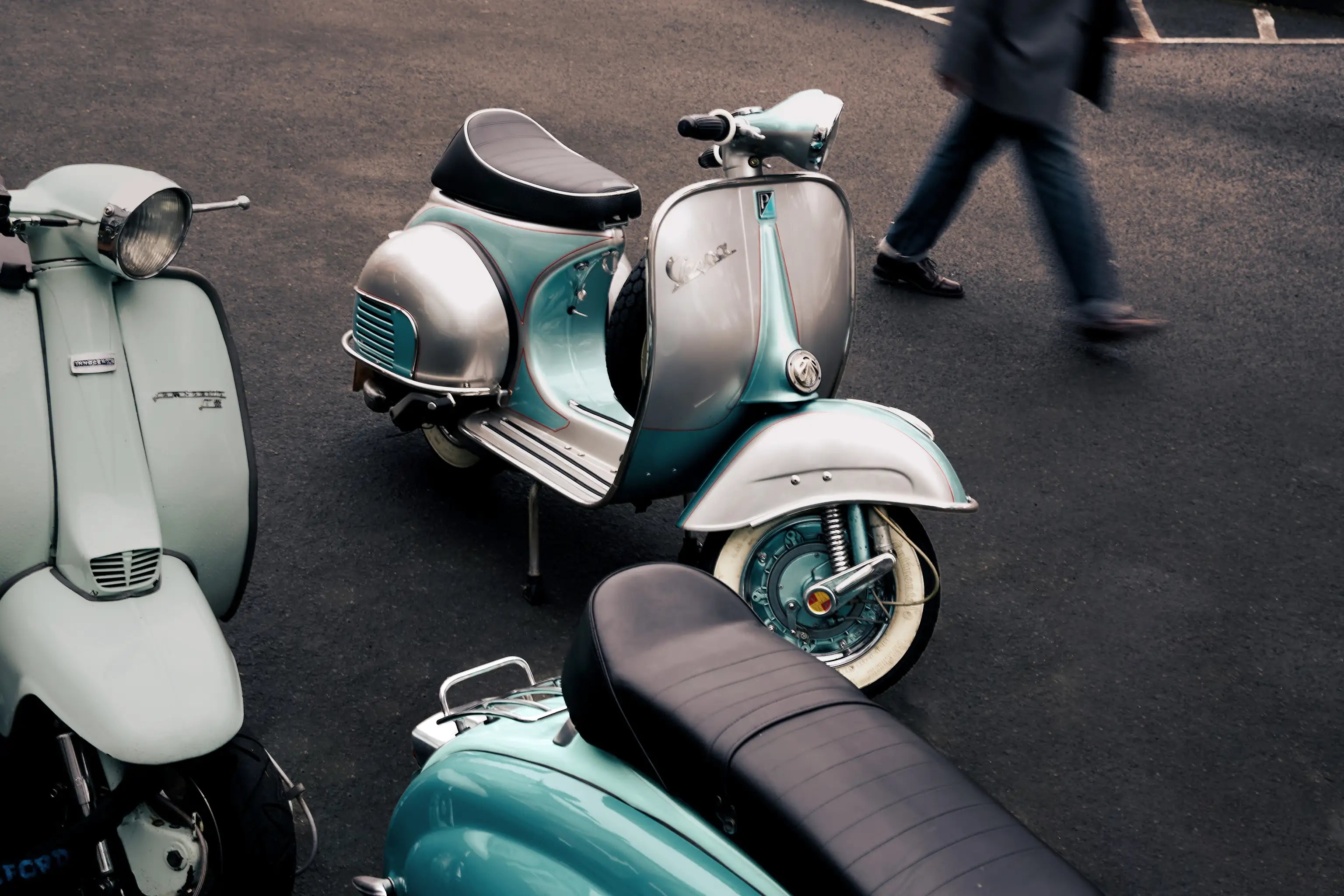 Two vintage-style scooters on a street with a person walking in the background.