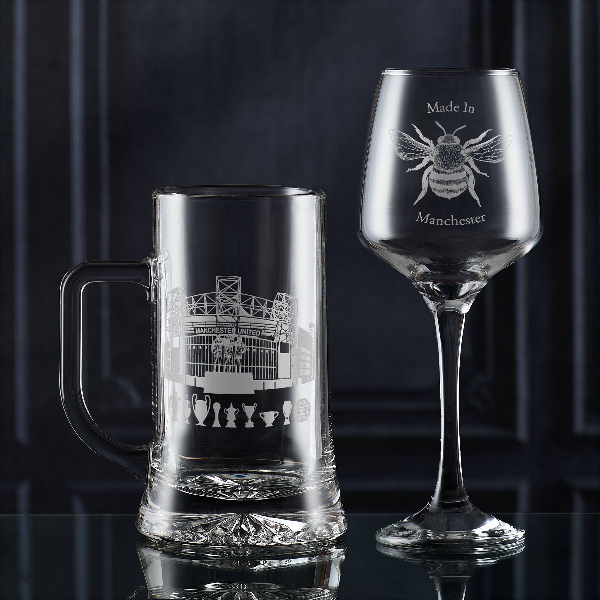 Clear glass mug and wine glass with engraved Manchester designs on a dark background