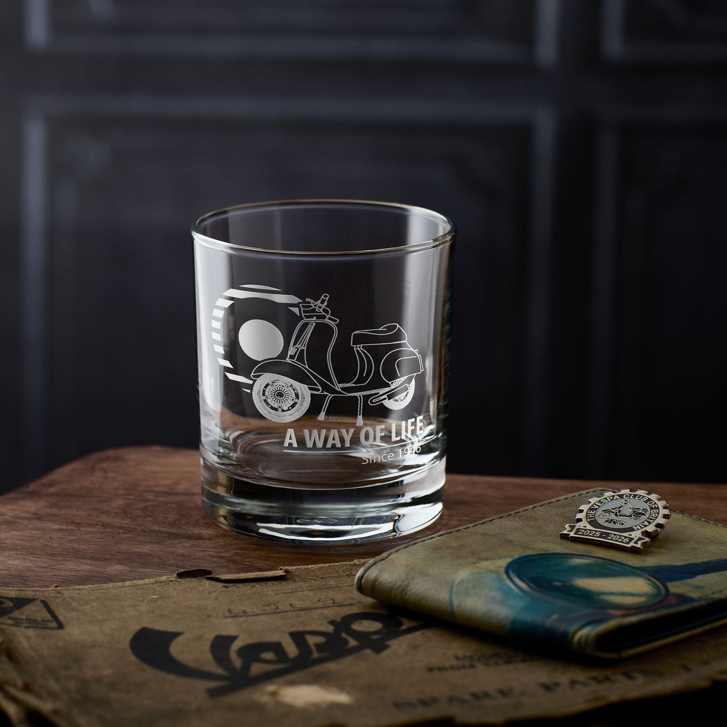 Whisky glass with a vespa 50 special scooter engraving on a wooden surface with The Vespa Club of Britain Badge.