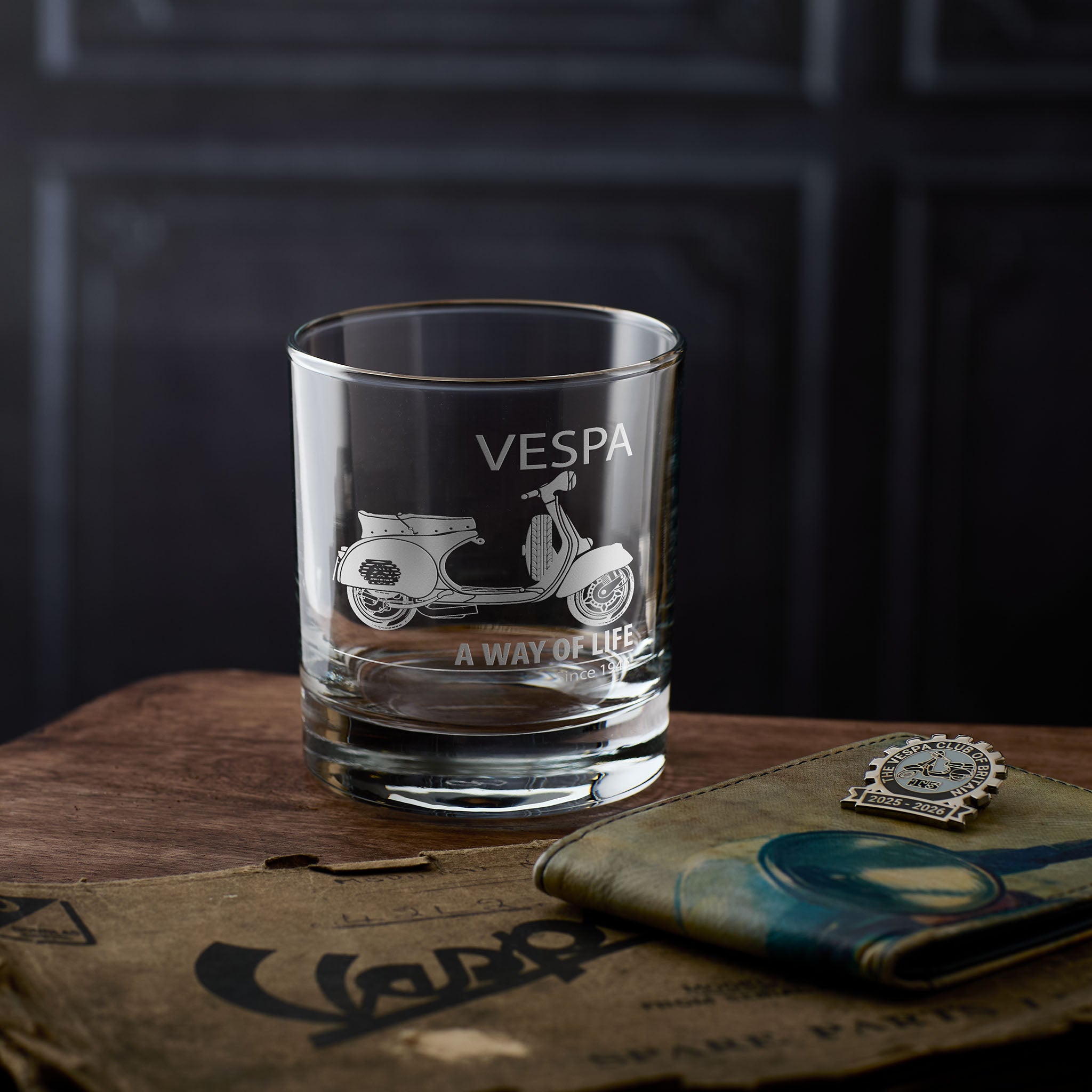 Vespa GS whisky glass with scooter design and text on a wooden surface with 42L2 booklet.
