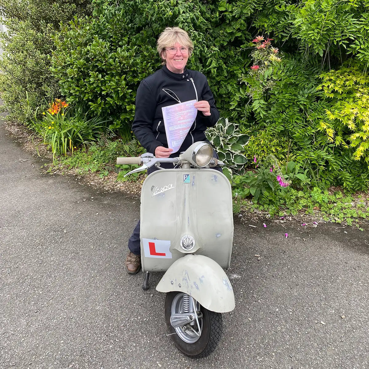 Person standing next to a scooter holding a document, with greenery in the background