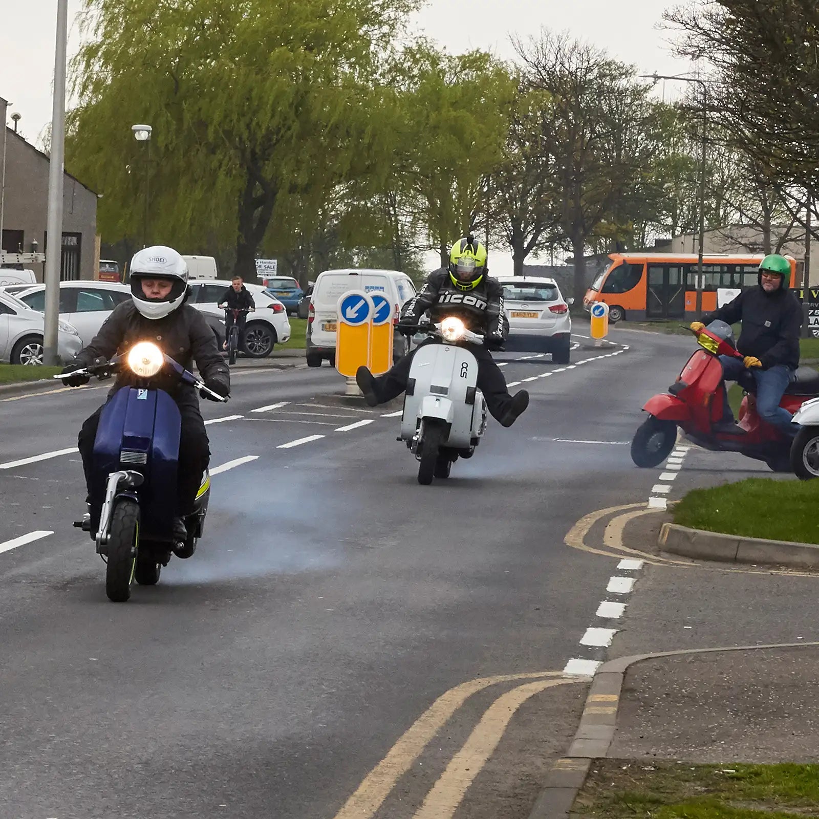 Three scooterists on a road with trees and vehicles in the background