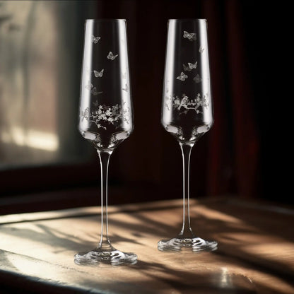 Two champagne flutes with engraved butterfly and. floral designs on a wooden surface.