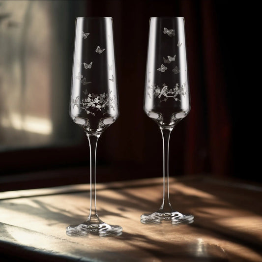 Two champagne flutes with engraved butterfly and. floral designs on a wooden surface.