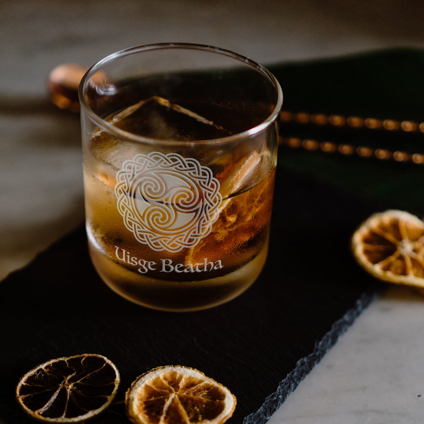 Glass of Scottish whisky with engraved water of life brand logo on a dark surface with dried orange slices.