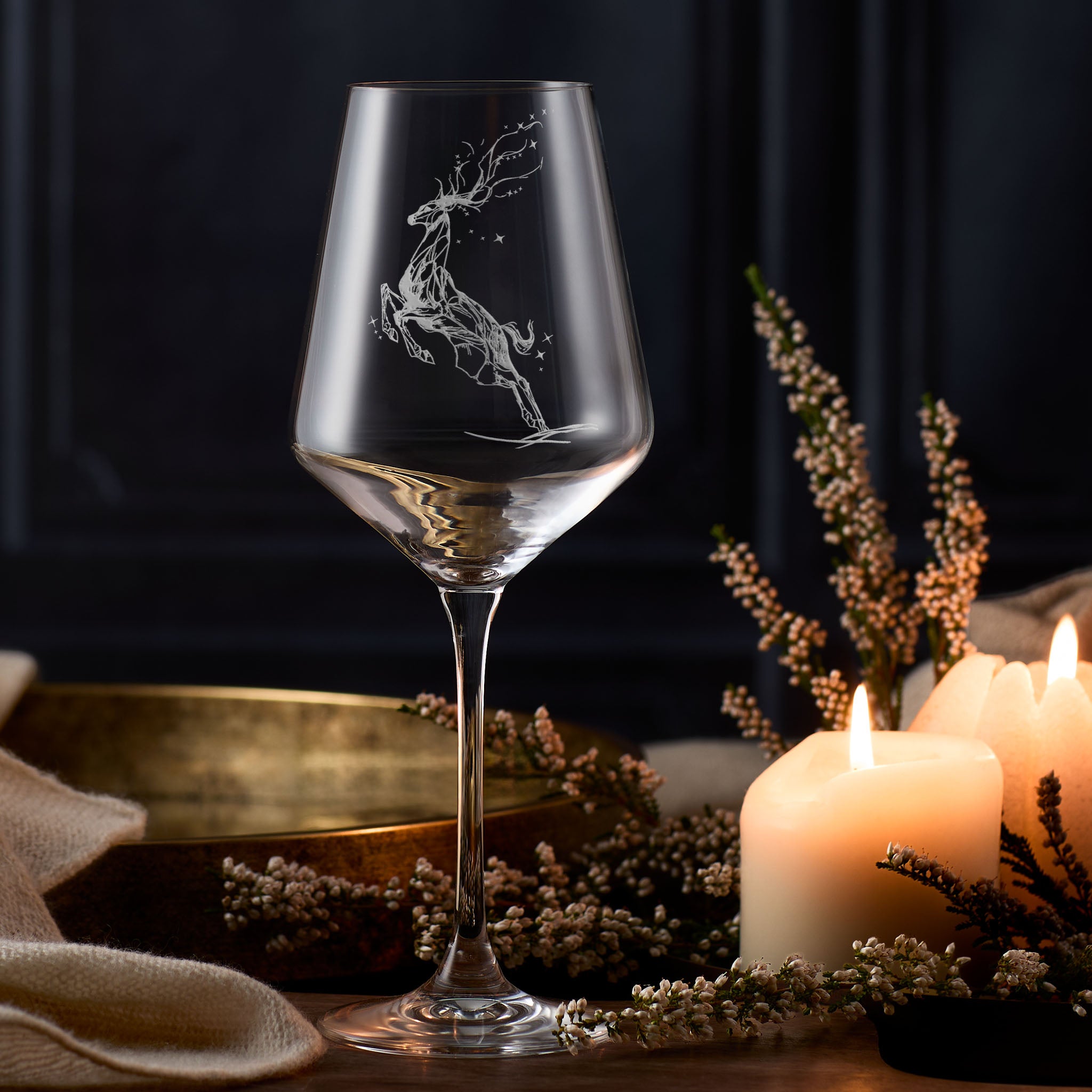 Wine glass with a deer leaping through the stars engraving on a dark background with candles and flowers