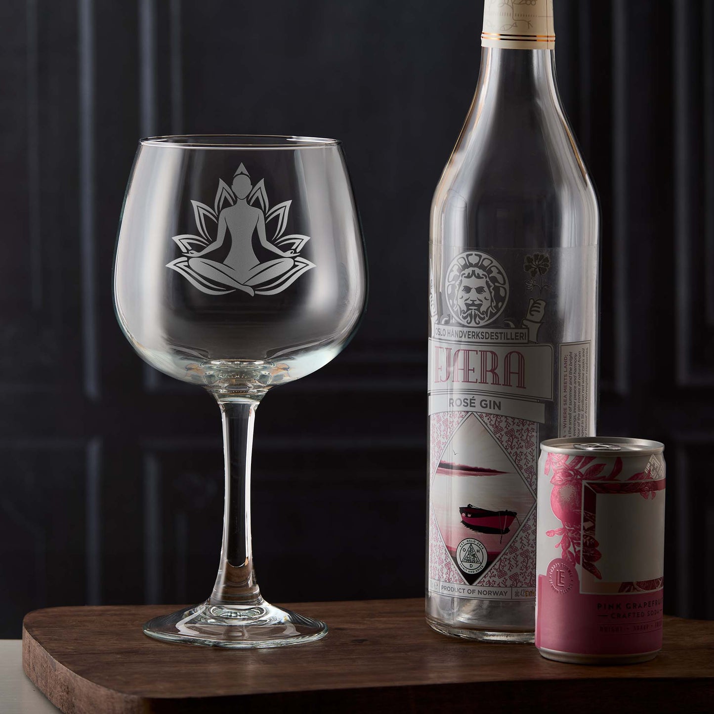 Gin glass engraved with a Yoga meditation designl, bottle of gin, and can on a wooden surface with dark background